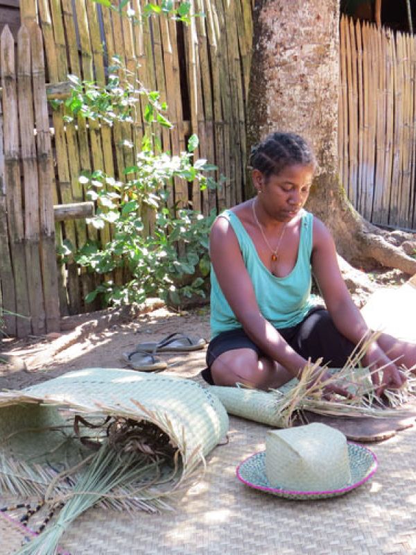 Artisan sitting on a plaited carpet, plaits baskets and has already plaited a hat.